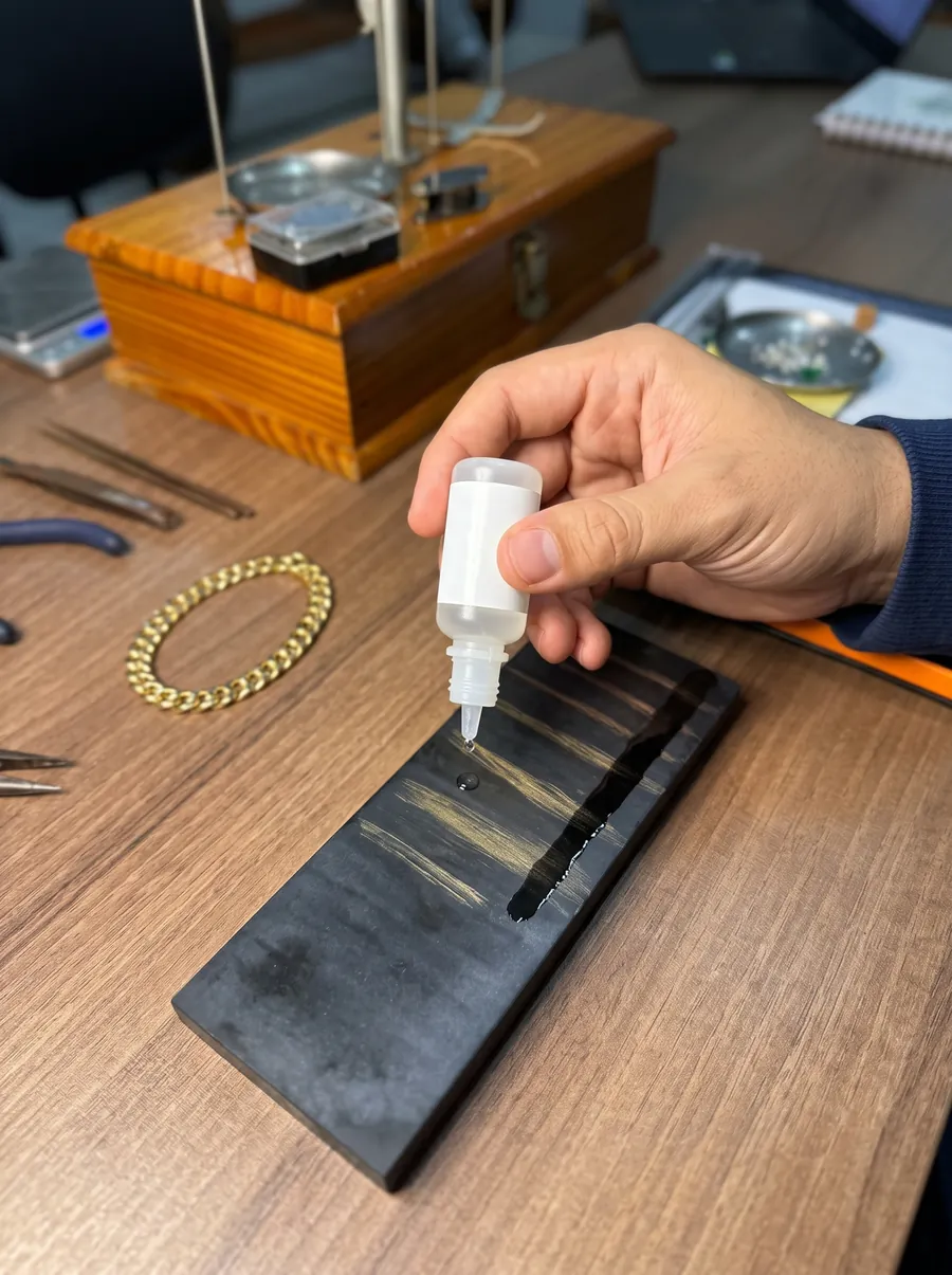 Avaliador pingando o líquido de teste sobre a pedra de toque riscada com ouro, ao lado de uma pulseira de ouro